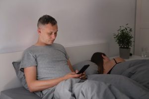 גבר שוכב במיטה עם הפלאפון ליד אישה ישנה